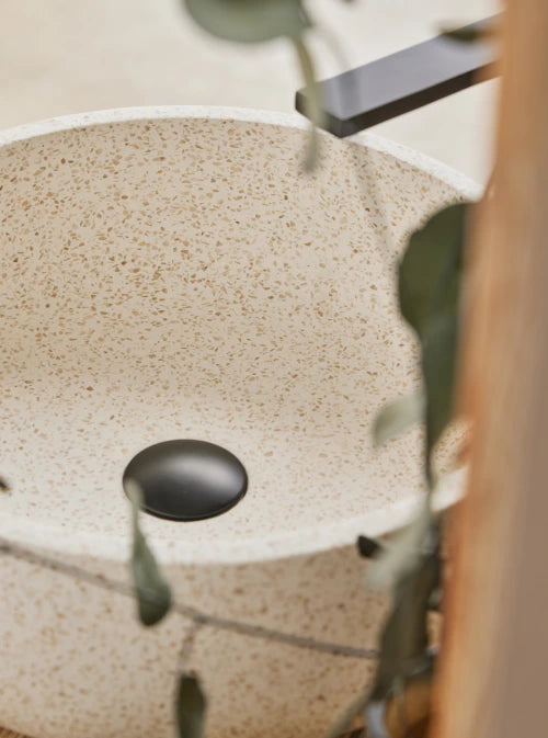 Beige, witte terrazzo waskom 39x39x13cm in close-up met subtiel gespikkeld patroon en zwarte plug, sfeervol gefotografeerd met groene bladeren.