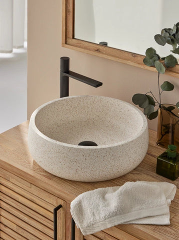 Ronde beige,witte terrazzo waskom 39x39x13cm met fijne korrel op houten badmeubel met zwarte kraan en natuurlijke accessoires in lichte badkamer.
