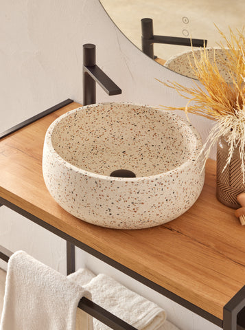 Witte, beige terrazzo waskom 39x39x14cm met kleurrijke chips in ronde vorm op licht houten blad, gecombineerd met zwarte opbouwkraan en decoratieve styling.