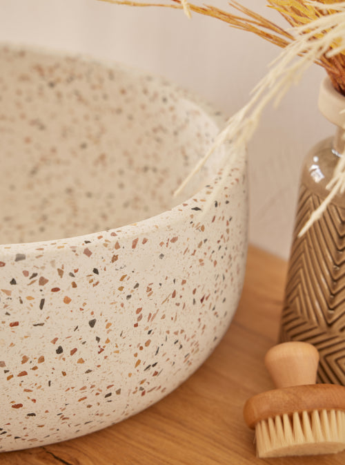 Witte, beige terrazzo waskom 39x39x14cm met kleurrijke chips in ronde vorm op licht houten blad, gecombineerd met zwarte opbouwkraan en decoratieve styling.