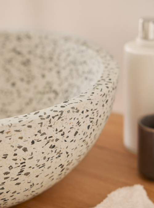 Ronde grijze terrazzo waskom 35x35x12cm met fijne korrel op houten badmeubel met zwarte kraan en natuurlijke accessoires in lichte badkamer.