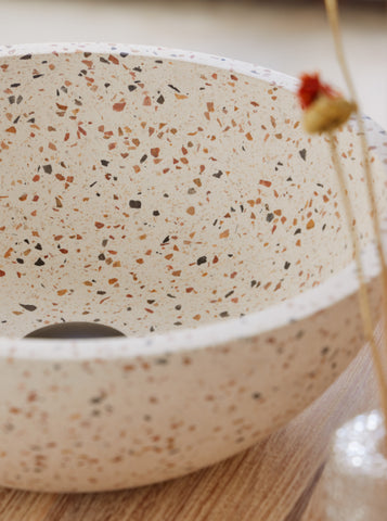 Ronde terrazzo waskom met multicolor steentjes op houten badmeubel met zwarte kraan en warme natuurlijke badkamerstyling 35x35x12cm