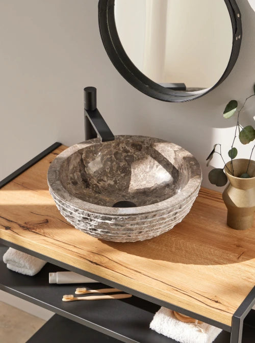 Natuursteen waskom in grijze tint op modern houten badmeubel met zwarte opbouwkraan en natuurlijke badkameraccessoires.