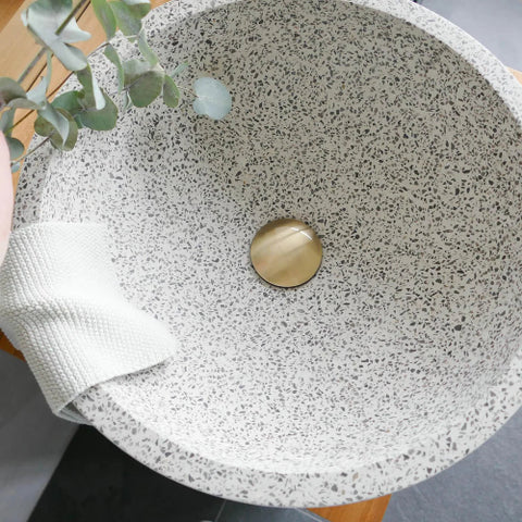 Binnenzijde van ronde wit grijze terrazzo gespikkelde natuursteen waskom met gouden afvoerplug en lichte handdoek